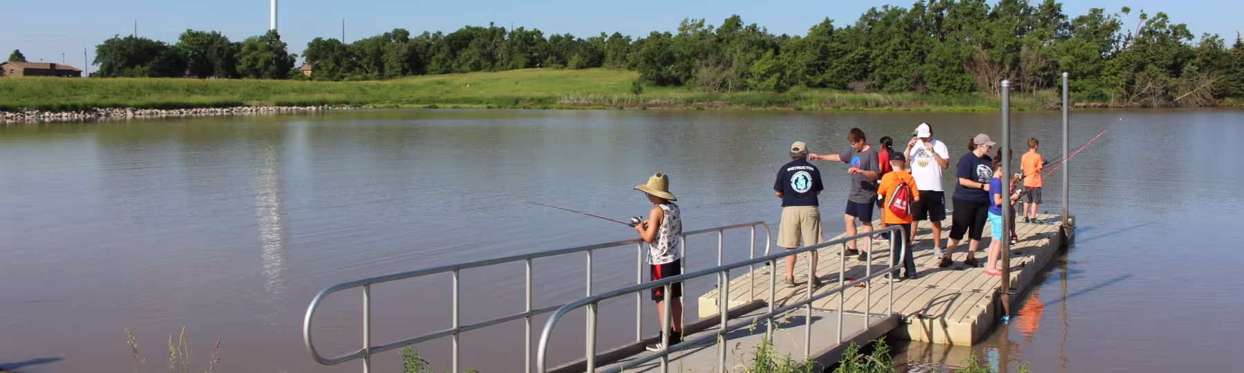 adults and children fishing off the dock at Recharge Lake; tree shoreline and watertower in the background